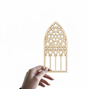 May include: A wooden cutout of a gothic window with intricate floral and geometric designs. The window features a pointed arch and multiple vertical panels. The natural wood color provides a neutral tone, suitable for various craft projects or home decor.