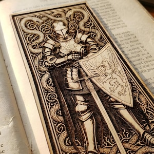 May include: A wooden bookmark featuring a detailed engraving of a knight in full armor, holding a sword and shield with a heraldic design. The bookmark is set against the backdrop of an open book, with the text "SVG" and "PNG" visible.