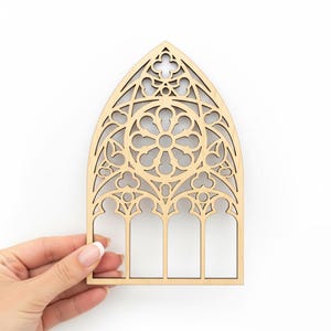 May include: A laser-cut wooden decorative piece in the shape of a cathedral window. The window features intricate geometric patterns and a central floral design. The wood has a natural, light brown color, and the piece is held by a hand against a white background.