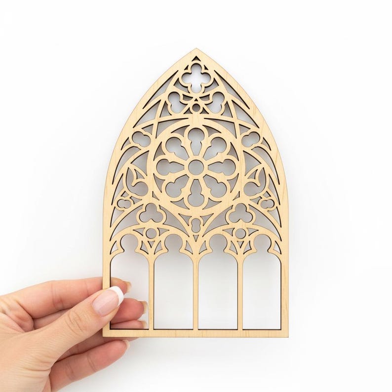 May include: A laser-cut wooden decorative piece in the shape of a cathedral window. The window features intricate geometric patterns and a central floral design. The wood has a natural, light brown color, and the piece is held by a hand against a white background.