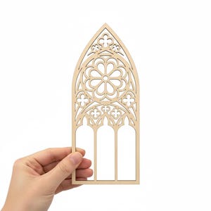 May include: A laser-cut wooden gothic window design. The window features a central floral pattern, trefoil details, and three arched panels. The natural wood color provides a neutral base for crafting or decoration. The window is held by a hand.