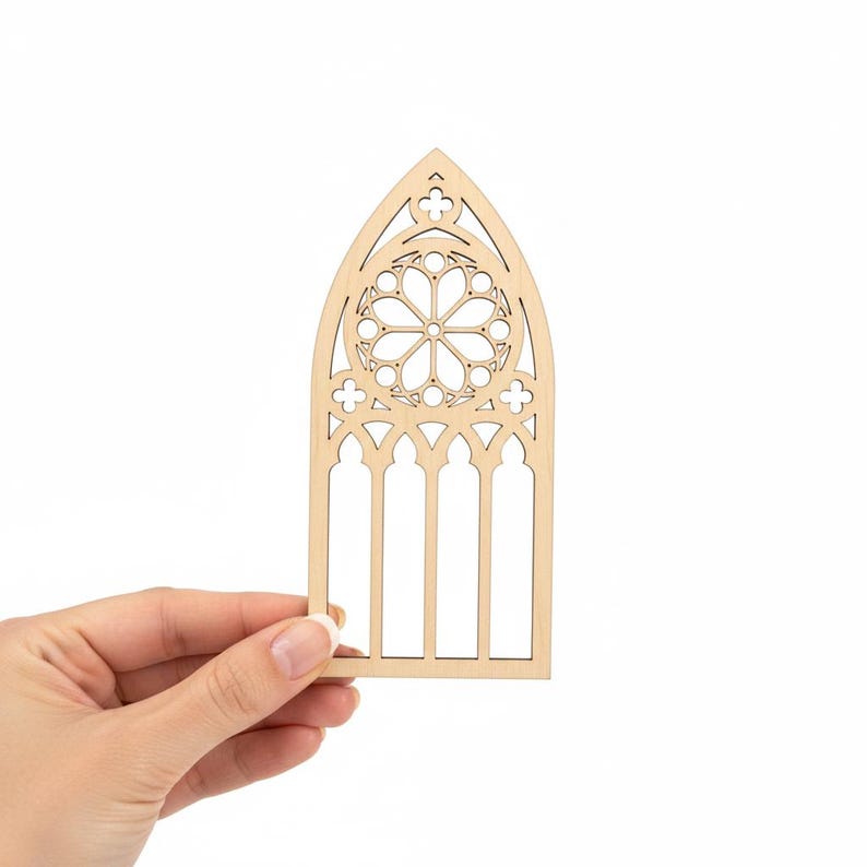 May include: A laser-cut wooden cathedral window design. The window features a rose window at the top and vertical rectangular openings below. The wood is a light tan color. The design is held by a hand against a white background.