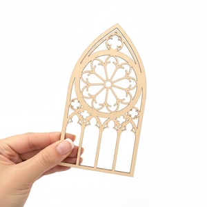 May include: A laser-cut wooden gothic window design. The window features a central circular rose window with radiating petal-like elements, surrounded by arched panels and decorative trefoil shapes. The wood has a natural, light brown color.