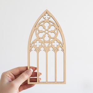 May include: A light-colored wooden cutout in the shape of a Gothic cathedral window. The window features intricate cut-out designs, including a central circular pattern and arched top. The cutout is held by a hand against a white background.