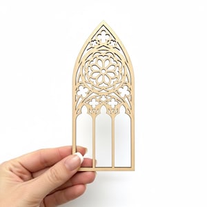 May include: A laser-cut wooden window frame with an arched top and intricate floral and geometric designs. The frame features three vertical rectangular openings and is held against a white background.