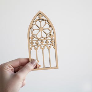 May include: A laser-cut wooden gothic window design. The window features a central circular pattern and arched openings. The natural wood color provides a neutral tone, suitable for various craft projects or decorative uses.