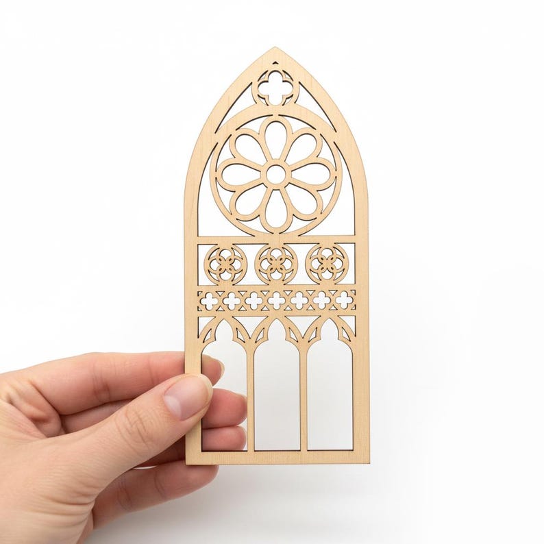 May include: A wooden cutout of a cathedral window, held by a hand. The window features a floral design at the top, three circular patterns, and a row of crosses. The cutout is made of light-colored wood.