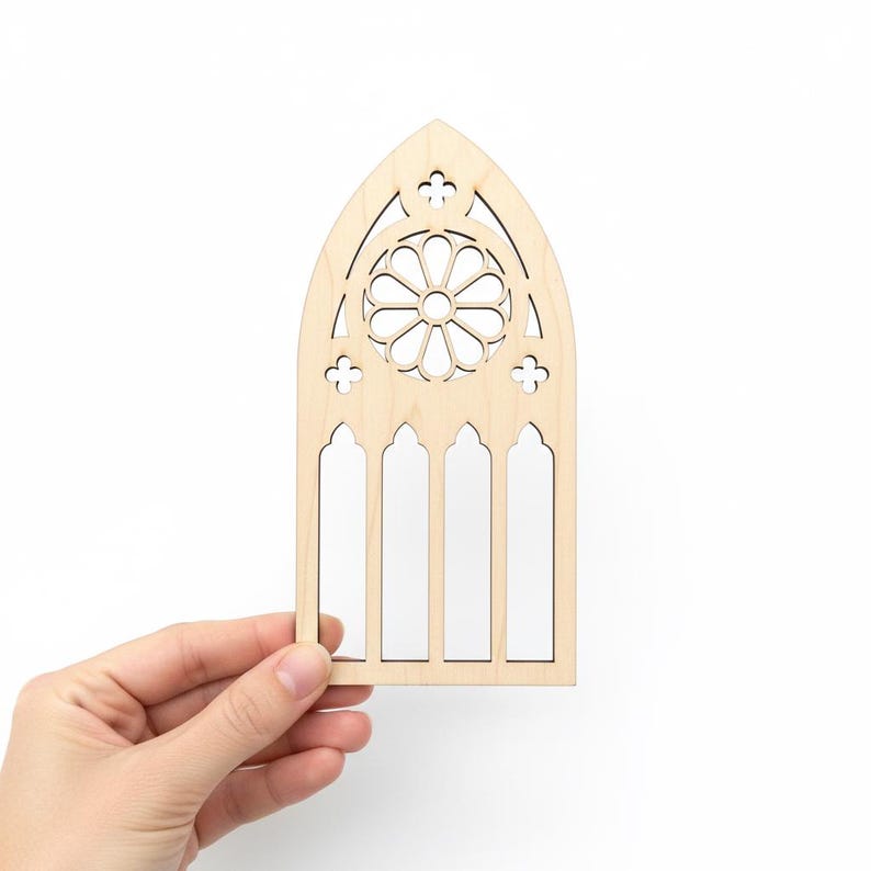May include: A laser-cut wooden gothic window design. The window features a central circular pattern and four arched window panes. The wood has a natural, light color. The design is held by a hand against a white background.