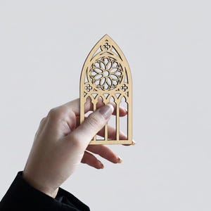 May include: A laser-cut wooden gothic window design with intricate floral patterns. The window is held by a hand against a plain background. The window is made of light-colored wood.