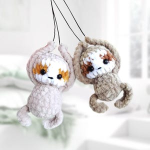May include: Two handmade crochet sloths hanging from black strings. The sloths have light brown and gray bodies, white faces with brown and orange markings, and black eyes. They are designed to look like they are hanging upside down.