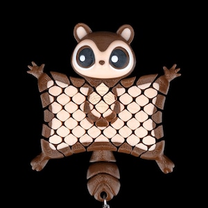 Könnte beinhalten: Ein brauner und beigefarbener 3D-gedruckter Flug-Eichhörnchen-Schlüsselanhänger. Der Schlüsselanhänger hat ein detailliertes Design eines Eichhörnchens mit ausgestreckten Armen und einem gemusterten Körper. Der Schlüsselanhänger ist an einer silbernen Kette befestigt.