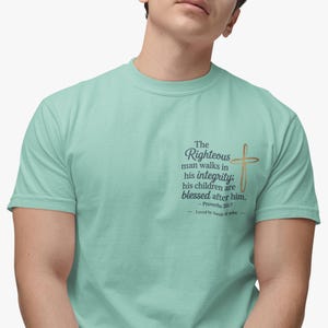 Puede incluir: Una camiseta azul claro con una cita sobre la justicia y una cruz dorada. La cita dice "El hombre justo anda en su integridad; sus hijos son bendecidos después de él. - Proverbios 20:7 Amado por Sarah af Job"