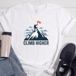 May include: White t-shirt featuring a mountain graphic with a person holding a flag at the summit. The text "CLIMB HIGHER" is below the mountain illustration. A travel mug, jeans, and a sneaker are visible.