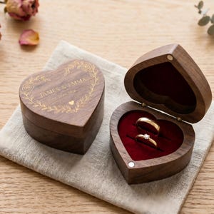 May include: Two heart-shaped wooden ring boxes, one closed and one open, displaying two rings. The closed box has the names "JAMES & EMMA" and a date engraved on the lid. The open box reveals a red velvet interior.