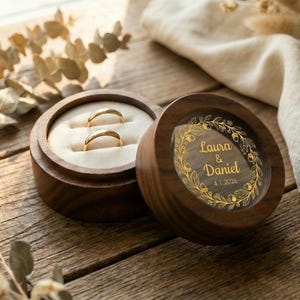 Pode incluir: Uma caixa de anéis de madeira com dois anéis de ouro dentro, ao lado da tampa aberta. A tampa tem os nomes "Laura & Daniel" e a data "4.1.2026" em letras douradas, rodeados por um desenho floral dourado. A caixa está sobre uma superfície de madeira.