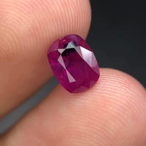 May include: A faceted, oval-shaped ruby gemstone. The gem is a deep, saturated red color with a polished surface. The ruby is held between two fingers, showcasing its brilliance and clarity. The background is dark.