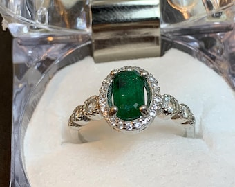 May include: A silver ring with an oval, emerald-green gemstone at the center, surrounded by a halo of smaller, clear gemstones. The ring is set in a clear plastic box with a white interior.