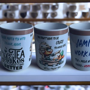 May include: Three white ceramic mugs with different designs. The first mug has a brown stripe and says "Tea Makes Everything Better". The second mug has a brown stripe and says "You don't have to be crazy to work here, we'll train you". The third mug has a brown stripe and says "Jamie's Work Mug Tea - Milk No Sugar Coffee - Never".