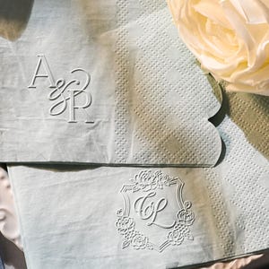 May include: Two pale blue napkins with scalloped edges, each embossed with initials. One napkin features "A&P" in an elegant script, the other "C&L" within a floral crest. A white rose and decorative plate complete the scene.