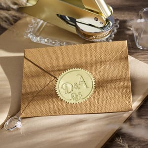 以下が含まれることがあります： イニシャル「D&A」と装飾的なデザインが施された円形のシールが付いた、金色の封筒。金のワックスシールスタンプとダイヤモンドリングも確認できます。封筒は薄茶色の表面に置かれています。
