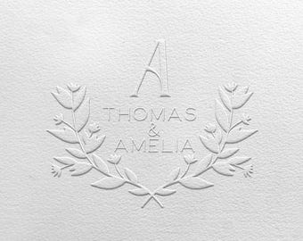 Könnte beinhalten: Ein weißes geprägtes Design auf strukturiertem Papier. Das Design zeigt die Namen "THOMAS & AMELIA" in einer Serifenschrift, umrahmt von einem floralen Kranz. Über den Namen steht ein elegantes "A".