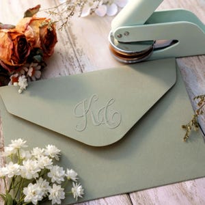 May include: A sage green envelope featuring an embossed monogram with the letters KD in a cursive script. A light mint green handheld embosser tool is positioned above the envelope, and small clusters of dried orange and white flowers are nearby.