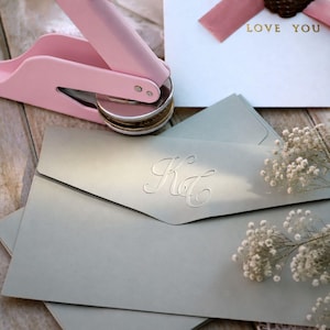 May include: A light pink handheld embosser tool positioned above sage green envelopes featuring a debossed monogram with the letters KC. A white card with a pink ribbon and the gold text LOVE YOU is visible in the upper right corner.
