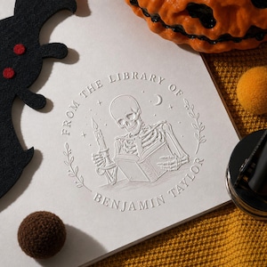 May include: A white paper surface featuring a circular embossed design of a skeleton reading a book while holding a candle. The text reads FROM THE LIBRARY OF BENJAMIN TAYLOR, surrounded by stars, a crescent moon, and a delicate leaf border.