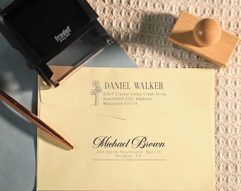May include: A light yellow envelope featuring two black ink return address stamps. The top stamp displays the name Daniel Walker with a floral graphic, and the bottom stamp shows the name Michael Brown. A black Trodat Printy 4913 stamp, a wooden handle, and a copper-colored pen are positioned nearby.