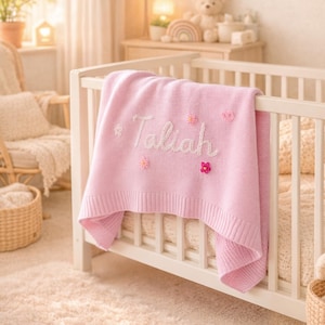 Op de afbeelding: Een zachtroze babydeken met de naam "Taliah" in witte letters geborduurd, versierd met bloemmotieven, over een witte wieg. De wieg staat in een kinderkamer, wat een zachte en warme sfeer creëert.