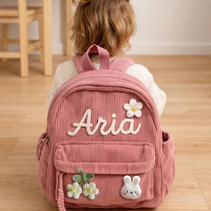 May include: Pink corduroy backpack with the name "Aria" embroidered in white script. The backpack features a front pocket, a small flower, and a bunny applique. A small child is wearing the backpack.