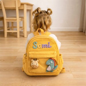 May include: A mustard yellow corduroy backpack with the name "Sami" embroidered in colorful thread. The backpack features a giraffe and dinosaur applique, side pockets, and adjustable straps. The backpack is designed for a child.