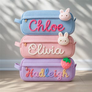 Puede incluir: Tres estuches de lápices de pana apilados. El superior es azul claro con el nombre "Chloe" en rosa. El del medio es rosa con el nombre "Olivia" en blanco. El inferior es morado con el nombre "Hadleigh" en colores del arcoíris.