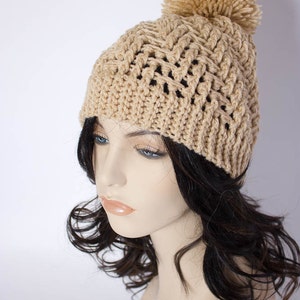 May include: A beige crocheted beanie with a fluffy pom pom on top. The beanie is worn on a mannequin with long, dark brown hair.