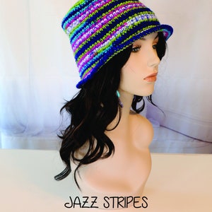 Peut inclure: Un chapeau crocheté rayé coloré dans des tons de bleu, vert, rose et blanc. Le chapeau a une large bordure et est posé sur une tête de mannequin. Le texte "JAZZ STRIPES" est écrit sous le chapeau.