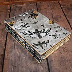 May include: A rectangular journal with a grey cover featuring a repeating pattern of black and white moths with occasional orange accents and black ink splatters. The book is bound with exposed black stitching along the spine and thick pages.