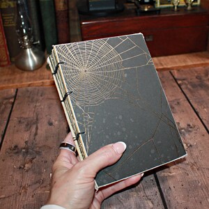 May include: A rectangular notebook with a matte black cover featuring a gold foil spider web design. The book has exposed stitching along the spine with black thread, visible paper edges, and a slim profile suitable for writing or sketching.