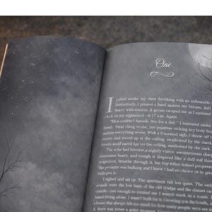 Open paperback book showing illustrated chapter page with moonlit sky design and grey interior pages under candlelight.