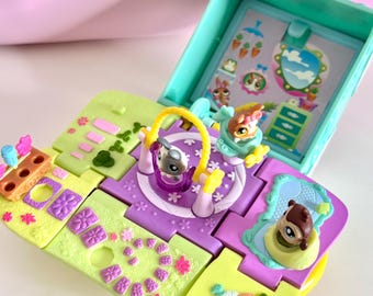 May include: A colorful, open toy playset with a teal box and multiple hinged sections. The sections feature various animal figures, floral designs, and a swing set. The background is a light pink.