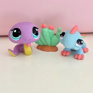 May include: A purple snake figurine with large eyes and a yellow belly, a green cactus figurine with pink accents on a brown base, and a light blue lizard figurine with a pink crest and feet, all featuring large, stylised eyes and smooth surfaces.
