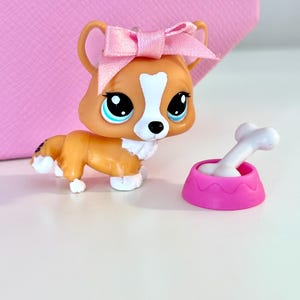 May include: A toy corgi dog figurine with a pink bow on its head and a pink food bowl with a white bone. The corgi is orange and white with large blue eyes. The food bowl is a bright pink color.