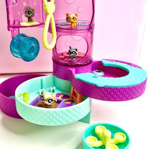 Puede incluir: Un colorido juego de juguetes LPS con múltiples niveles y compartimentos. El juego incluye pequeñas figuras de animales, una rueda de hámster azul y un clip amarillo. La estructura es principalmente morada, turquesa y rosa.