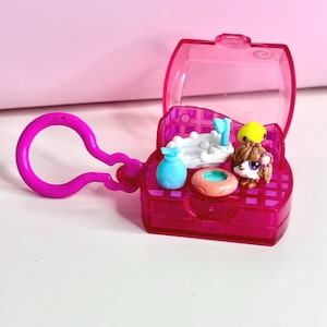 Puede incluir: Un mini juego rosa con tapa transparente, con una pequeña figura de perro, una bañera y otros accesorios. El juego incluye un llavero rosa para una fácil fijación. El diseño general es brillante y colorido, con un enfoque en los pequeños detalles.