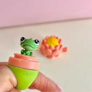Puede incluir: Un anillo de fantasía con una pequeña figura de rana verde sobre un diseño rosa y verde en capas. El anillo es de color verde brillante. Una flor rosa está en el fondo.