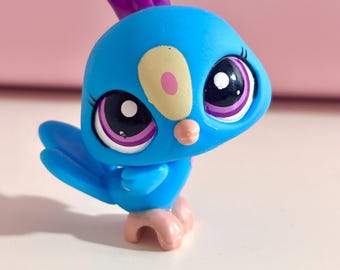 May include: A blue toy bird figurine with a purple crest and large, expressive eyes. The bird has a yellow and pink accent on its face and pink feet. The toy is designed with a cartoonish aesthetic.