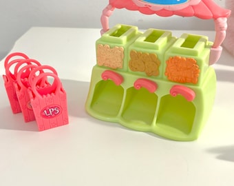 May include: A green and pink toy pet shop playset with three compartments, each with a different textured food design. A pink awning with a handle and a blue emblem tops the playset. Several small pink shopping bags with "LPS" text are to the left.