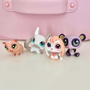 May include: Four colorful toy animal figurines. A light brown hamster, a white rabbit with a gray tail, a white and peach-colored bunny, and a purple panda with black ears. The toys are on a white surface with a pink background.