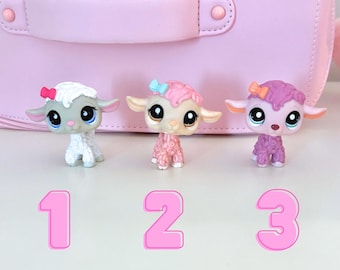 May include: Three small plush sheep toys with different colored fur. The sheep on the left is white with pink accents, the middle sheep is pink with light pink accents, and the sheep on the right is purple with pink accents. Each sheep has a bow on its head and large blue eyes.