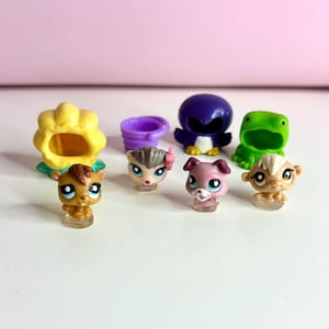 Puede incluir: Una colección de pequeñas figuras de animales coloridas. El surtido incluye una flor amarilla, un pingüino morado, una rana verde, un caballo marrón, un cubo morado, un erizo con un lazo rosa, un perro rosa y un mono beige.