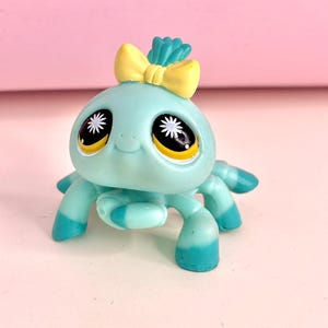 Puede incluir: Una araña de juguete azul claro con ojos grandes y un lazo amarillo. La araña tiene seis patas, cada una con un degradado de azul y turquesa. El juguete es de plástico y tiene una cara sonriente.